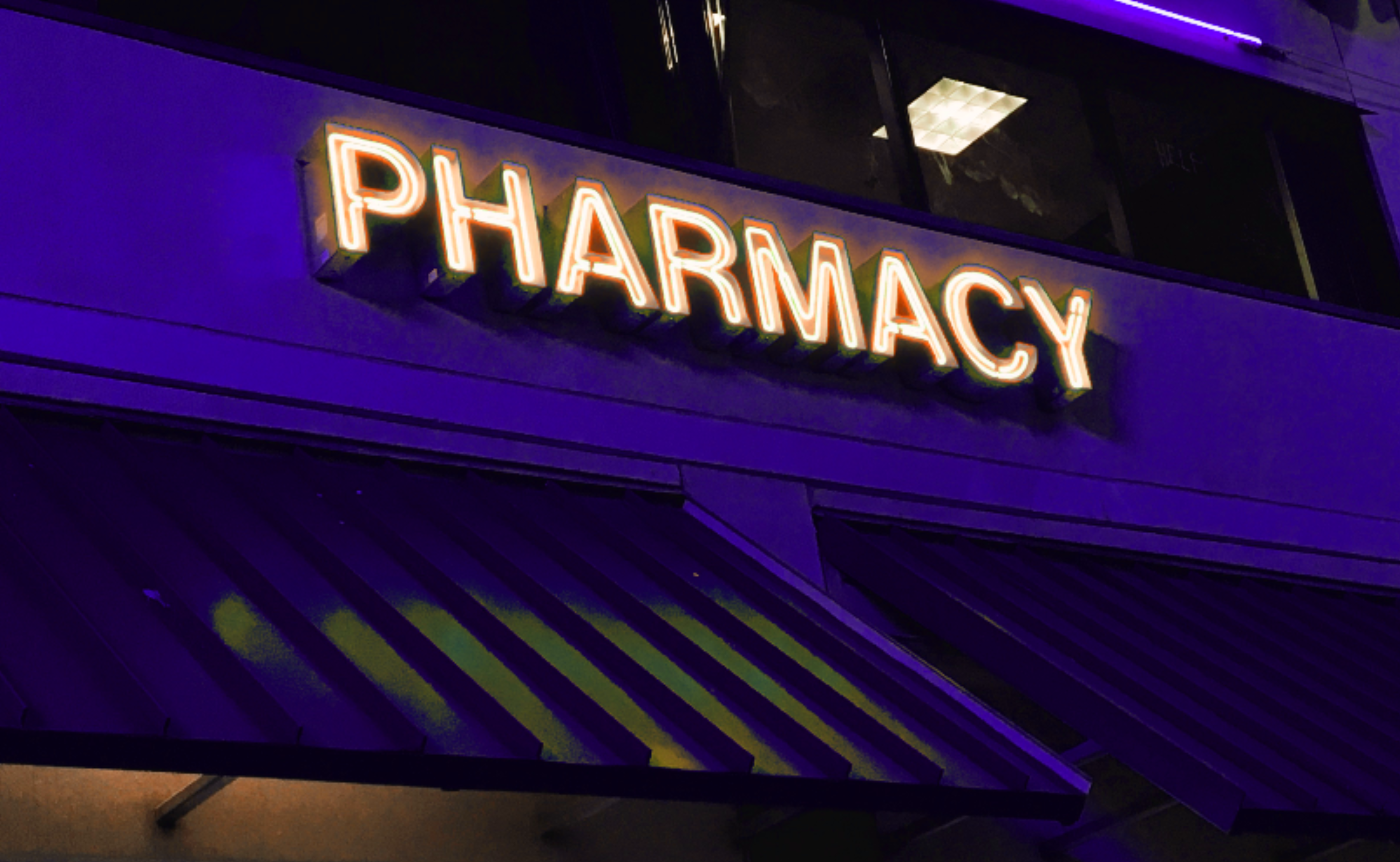 A glowing, orange pharmacy sign on an indigo building, with a purple light above the sign, and two dark awnings.