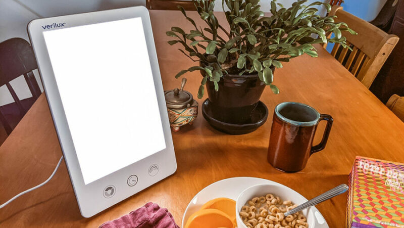 A POV of a light therapy lamp in front of a breakfast plate and coffee cup, on a wooden dining table.